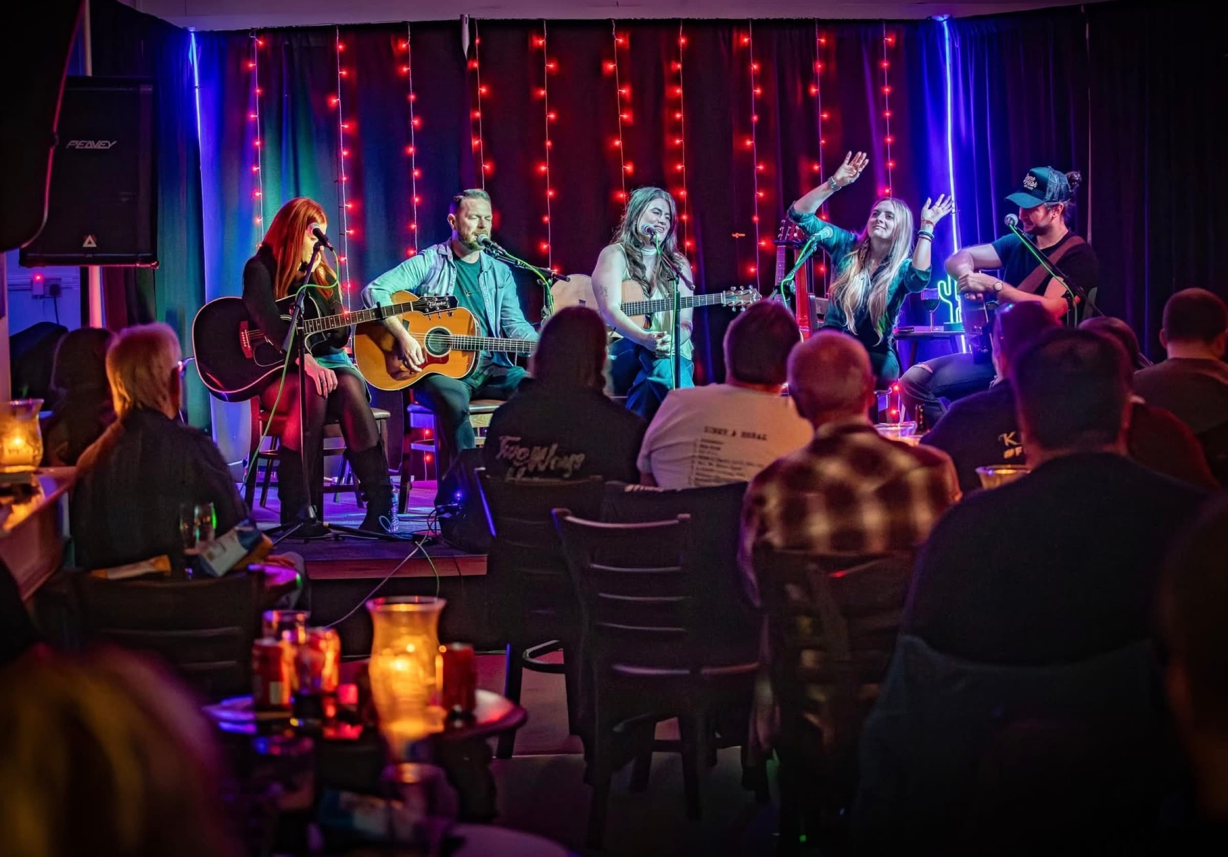 Katy Hurt's Good Company Songwriter's Circle: five musicians playing acoustic guitars in a row on the stage, the audience at candlelit tables