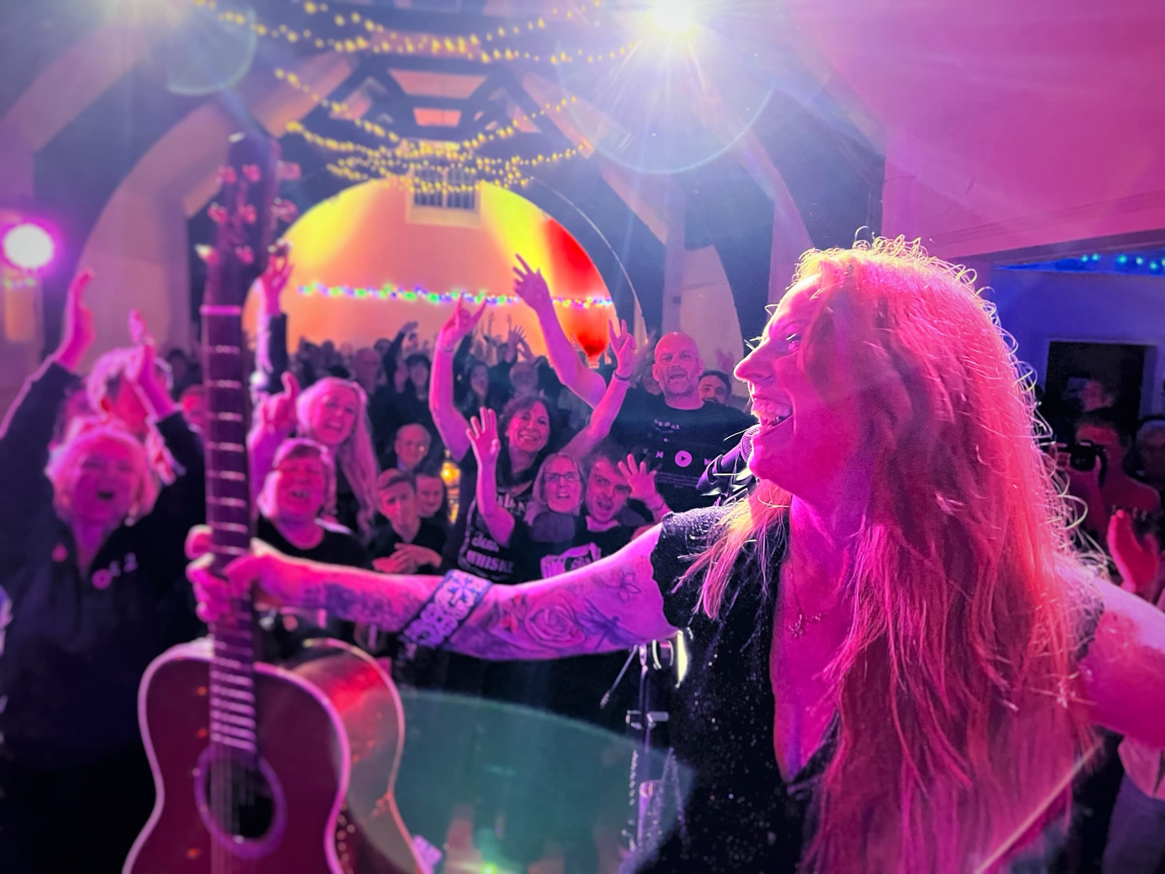 Kezia Gill: a singer with her guitar raised to a full room, arms up under the arched ceiling and fairy lights
